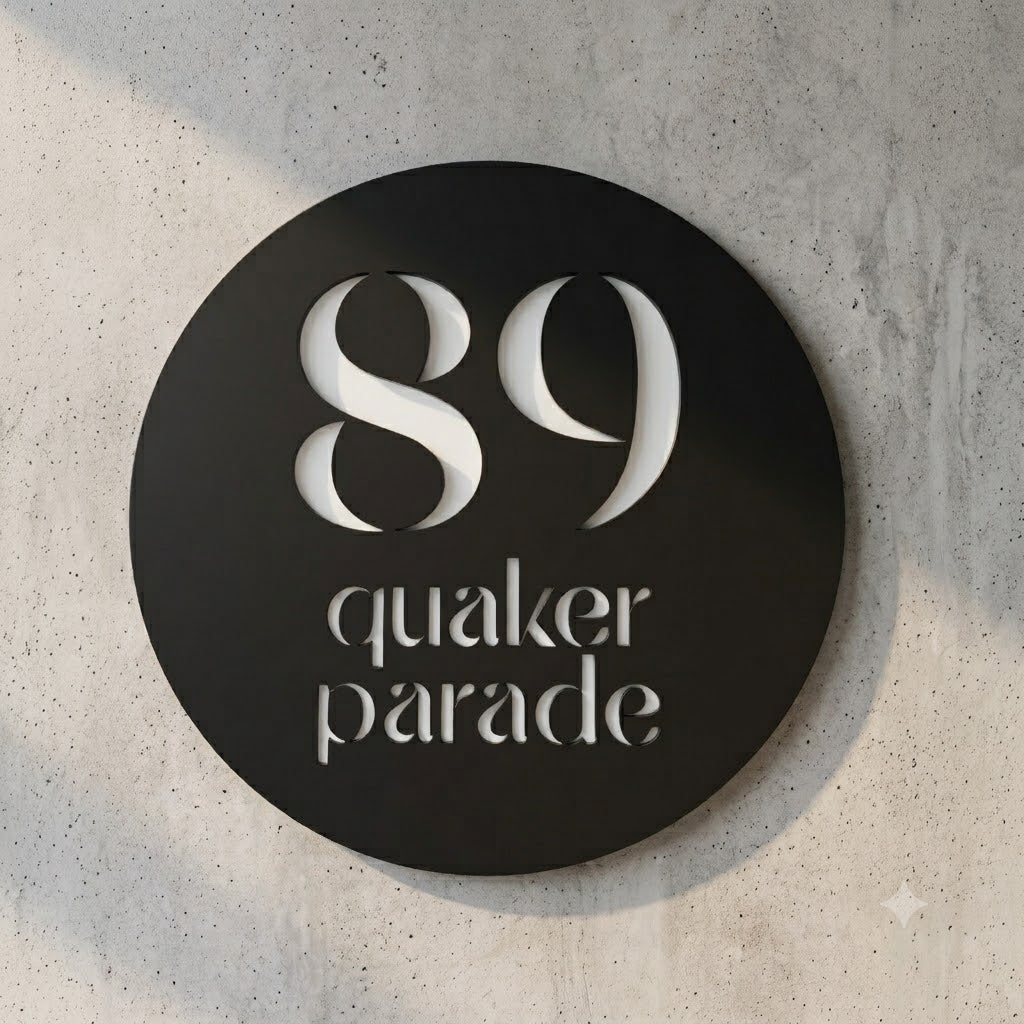A round black metal sign with white cutout lettering reading '89 quaker parade', mounted on a light gray textured concrete wall.