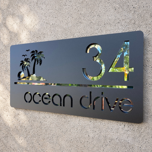 Metal house number sign with palm trees and '34 ocean drive' on a textured wall.